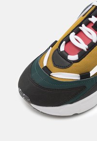 Nærbillede af tåen på en flerfarvet sneaker med sort, grøn, okker gul og rød stof, hvide snørebånd og hvid sål på grå baggrund.