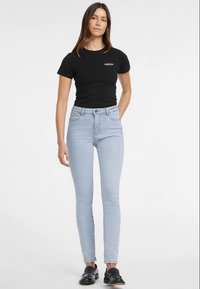 Mulher vestindo uma t-shirt justa preta, jeans skinny de cintura alta azul clara e sapatos loafers pretos, de pé contra um fundo branco.