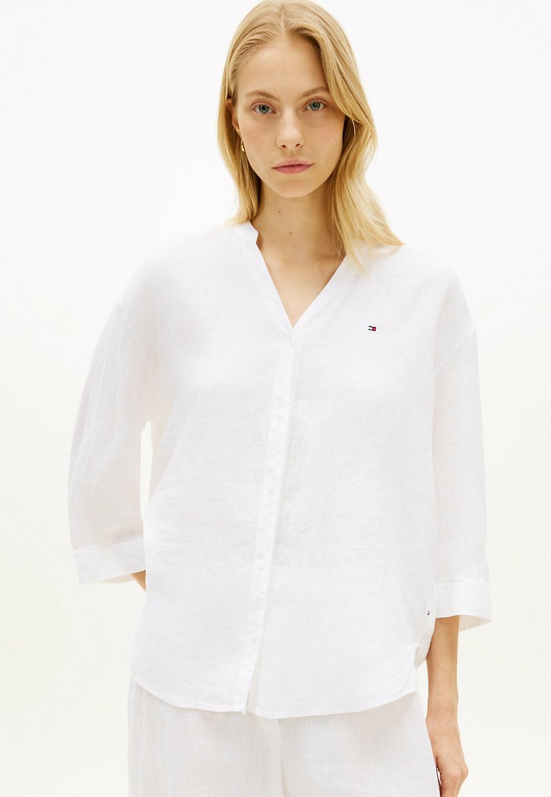Femme blonde portant une chemise blanche ample à boutons avec des manches trois-quarts et un pantalon blanc assorti, sur un fond uni.