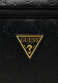 Black textured bag featuring embossed patterns and a triangular gold logo patch with "GUESS" and "WASHED JEANS" text detail.