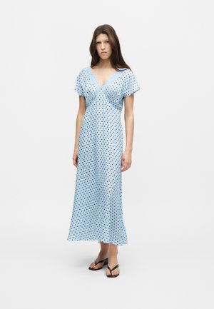 Femme aux longs cheveux bruns portant une robe longue bleu clair à manches courtes avec un col en V, à pois noirs, et des sandales tongs noires.