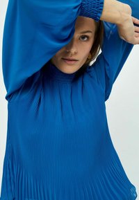 Blusa plissettata blu royal con maniche lunghe e fluide e colletto alto arricciato. Il tessuto ha una superficie liscia con morbidi drappeggi.