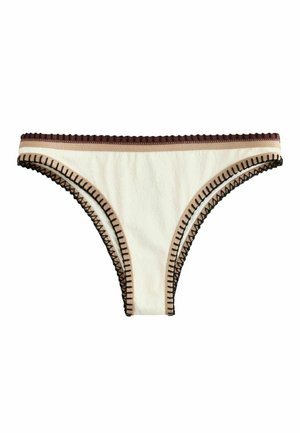 Femeie într-un bikini alb cu margini negre, pozând în fața unui perete texturat bej, purtând mai multe brățări cu mărgele, cu o mână în spatele capului.