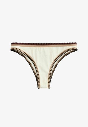 Femeie într-un bikini alb cu margini negre, pozând în fața unui perete texturat bej, purtând mai multe brățări cu mărgele, cu o mână în spatele capului.