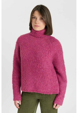 Vrouw met schouderlang bruin haar die een textuur roze coltrui en olijfgroene broek draagt tegen een effen achtergrond.