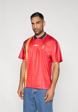 Czerwona koszulka polo z krótkim rękawem, z geometrycznym żółto-niebieskim wzorem z jednej strony, z prążkowanym granatowym kołnierzem i logo Adidas.