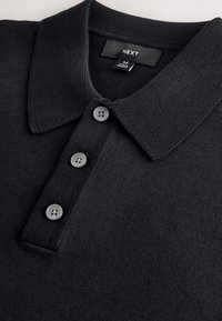 Polo noir en tissu tricoté. Il présente un col classique, quatre boutons gris, et une étiquette indiquant "NEXT" avec la taille M.