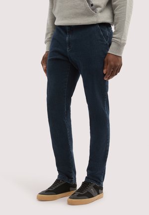 Hombre con jeans azul oscuro y zapatillas negras con suelas de goma, de pie con las manos relajadas a los lados contra un fondo liso.