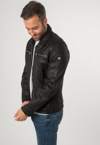 Giacca di pelle nera con una finitura testurizzata, dotata di zip frontale, due tasche sul petto e colletto standard. Indossata con jeans blu.