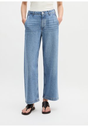 Persoon draagt lichtblauwe high-waist wijde pijpen jeans, witte top, zwarte sandalen, staat met handen in de zakken tegen een effen achtergrond.