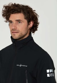 Svart jacka med hög krage och framdraget blixtlås, med en logotyp på bröstet och reflexdetaljer på vänster ärm. Slät textur.