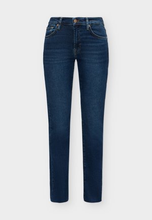 Femme portant une veste en cuir noire, un haut noir, un jean bleu, des chaussures noires pointues, tenant un sac à main noir à franges sur un fond blanc.