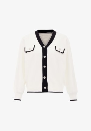 Valkoinen neuletakki mustilla yksityiskohdilla, V-kaula-aukko, kaksi rintataskua, koristeelliset napit ja sahalaitaiset taskun reunat. Rakenteellinen neulekangas.