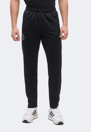 Uomo che indossa pantaloni da allenamento neri slim fit con elastico in vita e sneakers bianche con strisce nere, in piedi su sfondo chiaro.