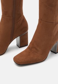 Bottines en daim marron avec un bout pointu, une fermeture éclair latérale et un talon bloc métallique. Texture lisse avec un design épuré.