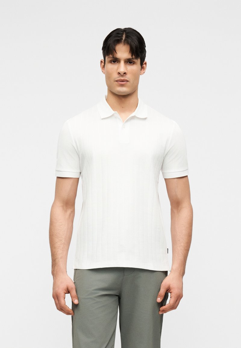 Homme debout, face à l'avant, portant un polo blanc à manches courtes et un pantalon gris, sur un fond clair uni.