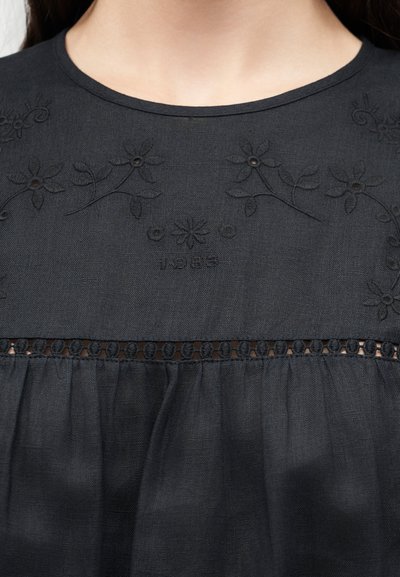 Μαύρο υφασμάτινο κορμάκι με κεντημένα λουλουδένια σχέδια και το έτος "1985" ραμμένο πάνω από οριζόντια δαντελωτή κοπή.