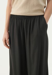 Pantalons noirs amples avec une taille élastique. Le tissu semble lisse et léger, atteignant le milieu du mollet, avec des poches latérales visibles.