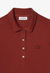 Maroon Lacoste-polo paita, jossa on kapea malli, klassinen kaulus, kolmen napin listä ja brodeerattu logo vasemmassa rinnassa. Teksturoitu kangas.