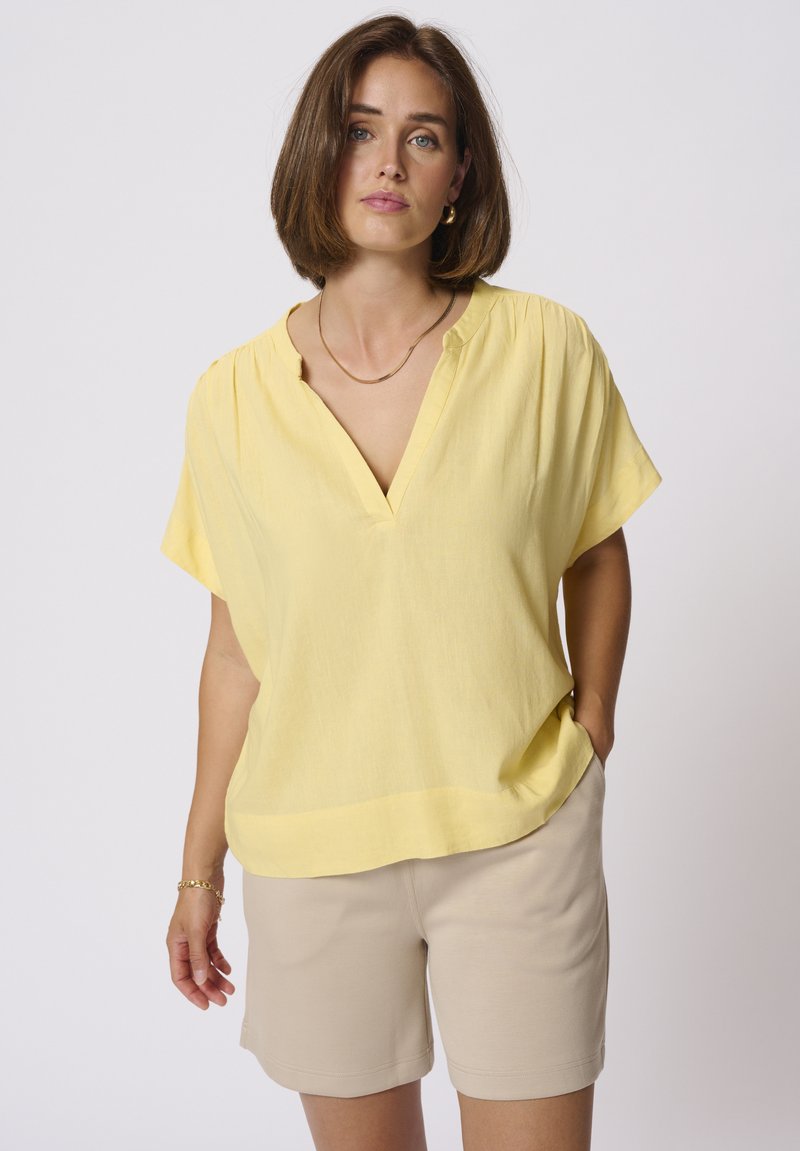 Blouse jaune à manches courtes avec un décolleté en V, en tissu texturé et à coupe décontractée. Associée à un short beige, doté de poches latérales.