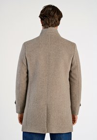 Mand iført en knælang, beige uldfrakke med rygsplit, der står med ryggen til mod en ensartet lys baggrund.