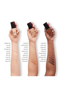 Drie armen tonen verschillende tinten foundation in glazen flessen met zwarte doppen. De swatches variëren van lichte tot diepe tinten, gelabeld ter referentie.