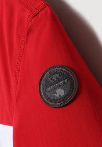 Rode stoffen jacksleeve met een zwarte cirkelvormige patch met oranje accenten, die een wereldbol en tekst toont met de aanduiding 'mapping sinds 1937'.