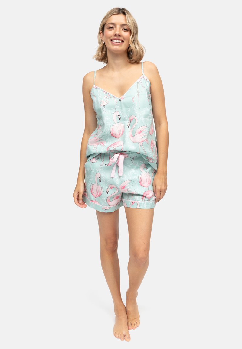 Junge Frau lächelt, trägt ein passendes hellblaues Camisole- und Shorts-Set mit pinkem Flamingo-Motiv, steht barfuß vor weißem Hintergrund.
