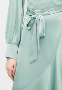 Αριστερό χέρι και χέρι σε μακρυμάνικο φόρεμα μέντας πράσινου χρώματος με μανσέτα στον καρπό, με ζώνη από ύφασμα δεμένη στη μέση.