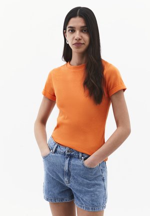Frau mit langen dunklen Haaren, die ein oranges kurzärmeliges Shirt und blaue Jeansshorts trägt, steht mit den Händen in den Taschen vor weißem Hintergrund.