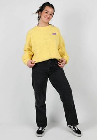 Pull en tricot jaune avec un motif en zigzag, associé à un jean noir taille haute et des baskets noires. Présente un petit écusson de logo violet.
