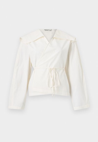 Λευκό τοπ με μακριά μανίκια που δένει μπροστά, με φαρδιά γιακά, παρουσιάζεται σε απλό φωτεινό φόντο.