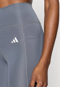 Γκρι κολάν από ελαστικό ύφασμα, με ψηλή μέση, πλαϊνή τσέπη και λευκό λογότυπο στο μηρό. Λείο ύφασμα και εφαρμοστή σχεδίαση.