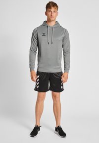 Grijze lange mouwen hoodie met trekkoord, zwarte shorts met witte chevronstrepen aan de zijkanten, en zwarte sportschoenen.