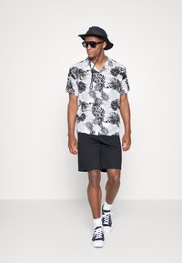 Short-sleeve black and white floral shirt, black shorts, black bucket hat, and sunglasses; walking on a plain background. Black sneakers with white soles.