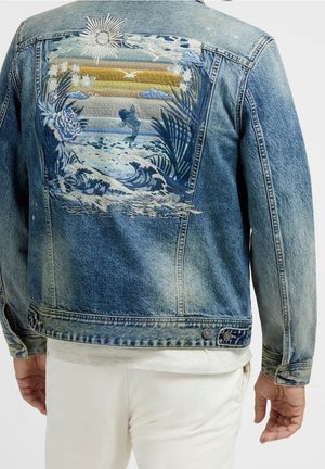 Chaqueta vaquera con bordado de atardecer tropical, sol, olas, peces, flores y hojas de palma en la espalda, combinada con pantalones blancos.