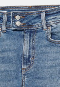 Blugi din denim albastru cu nasturi metalici dubli, bucle pentru curea, cusături vizibile și fermoar parțial deschis.