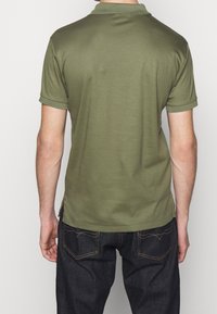 Olivgrön polo med korta ärmar, mjuk textur, platt stickad krage och sidoflikar, i kombination med mörka jeans.