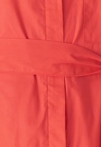 Tkanina v koralni barvi z gladko teksturo ima zavezano pasovno vrvico, navpične naborke in en sam žep na levi strani.