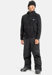 Zwart fleece sweatshirt met een hoge hals en logo; gecombineerd met loszittende zwarte sneeuwbroeken. Robuuste textuur en functioneel ontwerp.