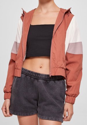 Kobieta ubrana w kolorową kurtkę przeciwwiatrową, czarny krótki top i wyblakłe czarne szorty z elastycznym pasem, widziana od szyi do połowy uda.