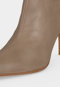 Gros plan d'une bottine en cuir taupe avec un bout pointu et un talon en bois empilé sur fond blanc.
