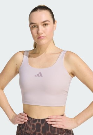 Jonge vrouw met lichte huid en haar naar achteren gebonden draagt een lichtpaarse sportbeha en legging met luipaardprint, staand met handen in de zij.