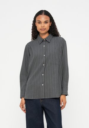 Camicia grigia a righe con bottoni, colletto, maniche lunghe e taschino sul petto. Presenta righe verticali bianche su un tessuto texturizzato.