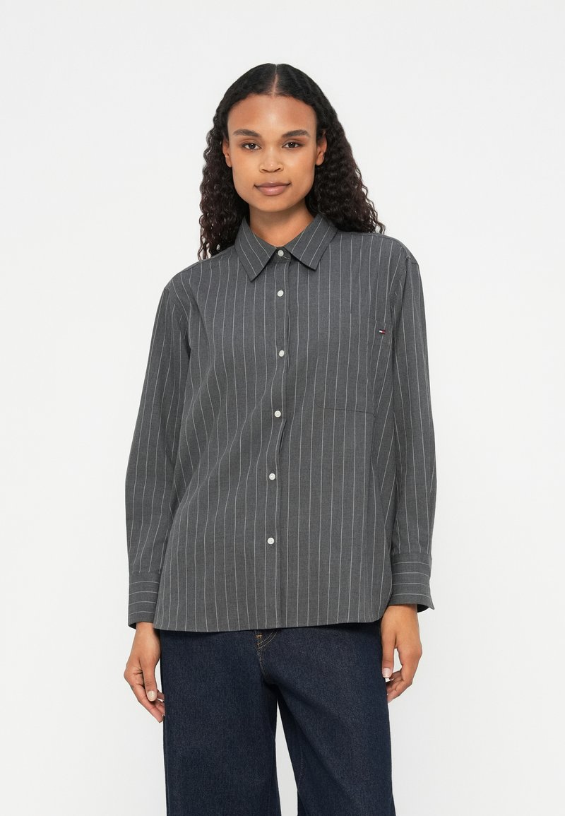 Camicia grigia a righe con bottoni, colletto, maniche lunghe e taschino sul petto. Presenta righe verticali bianche su un tessuto texturizzato.