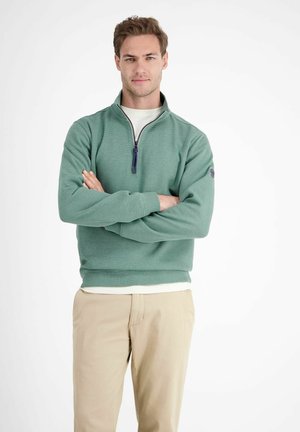 Grüner Pullover mit halbem Reißverschluss, strukturiertem Stoff, Rippbündchen und hohem Kragen. Getragen über einem weißen Hemd, kombiniert mit beigen Hosen.