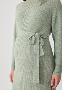 Rochie tricotată verde cu un design texturat din nervuri, având un cordon la talie și mâneci lungi. Fabricată dintr-un material moale și confortabil.