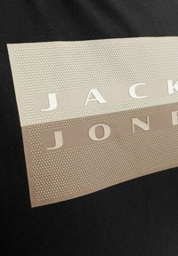 Svart T-skjorte med teksturert logo i beige, med hevede prikker og bokstaver som staver "JACK & JONES" over designet.