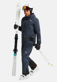 Giacca e pantaloni da sci di colore blu scuro, scarponi da sci bianchi, in mano sci e bastoncini. Presenta cerniere e tessuto testurizzato, con accenti neri a contrasto.