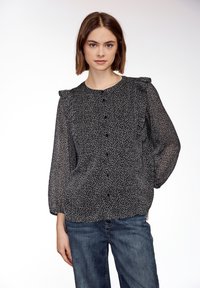 Jeune femme aux cheveux bruns mi-longs, portant un chemisier noir et blanc à motifs avec des épaules à volants et un jean bleu, debout devant un fond blanc.